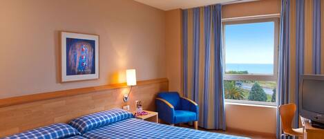 Habitación de hotel con dos camas, un escritorio con televisor, una silla, una lámpara y una ventana con cortinas.