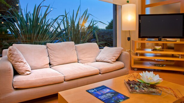 Een moderne woonkamer met een beige bank, een bijzettafeltje met een tijdschrift, een tv op een houten dressoir, en een groot raam met uitzicht op hoge planten en een heldere lucht.