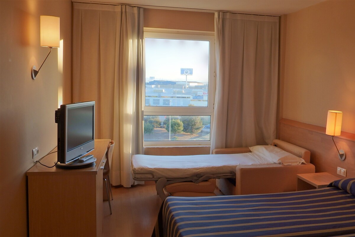 Habitación de hotel con cama, escritorio con computadora, ventana con cortinas y un cuadro en la pared.