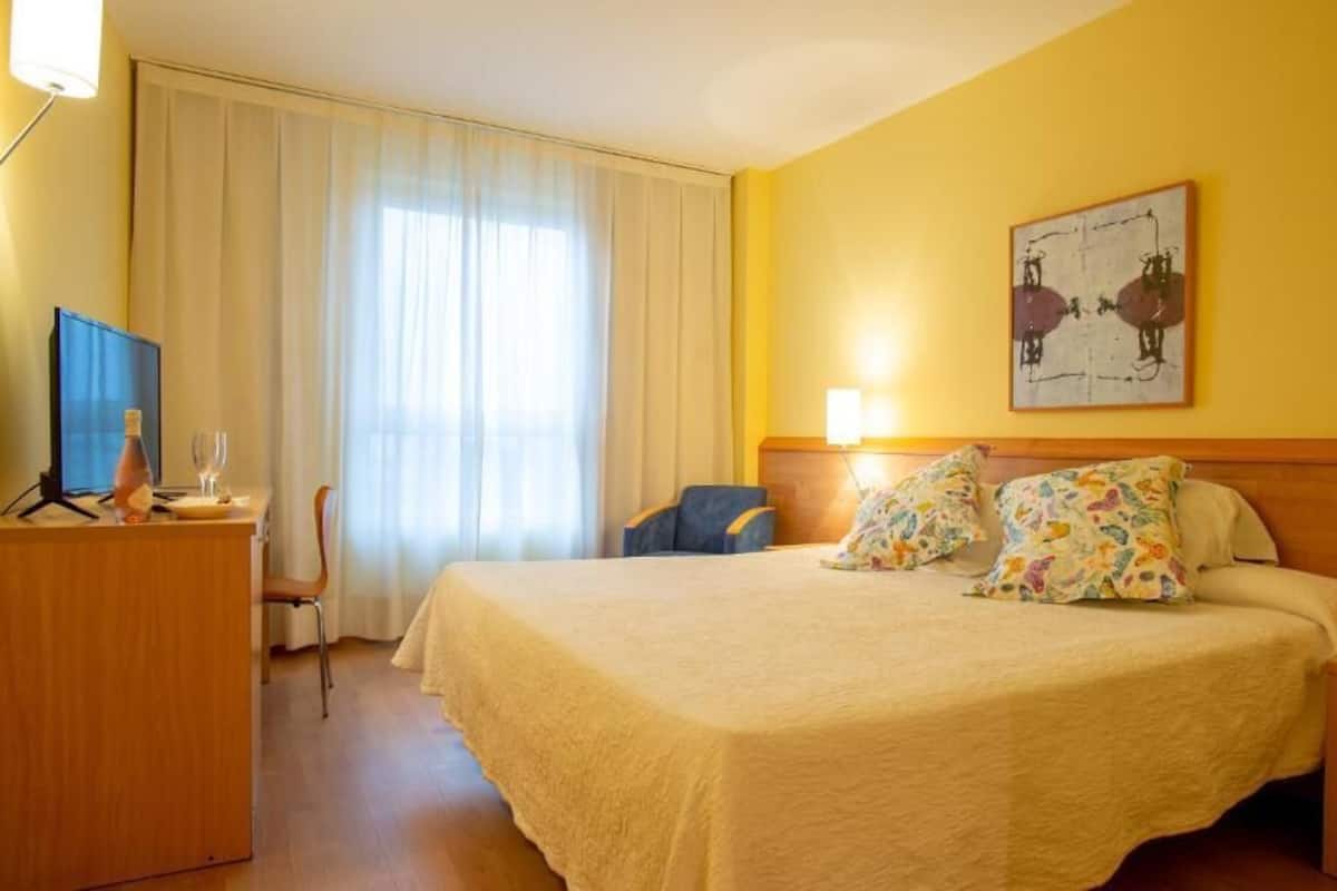 Habitación de hotel con cama, escritorio, silla, televisor y ventana con cortinas.