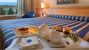 Een hotelkamer met een bed, een eettafel met ontbijtartikelen, uitzicht op zee en een schilderij aan de muur.