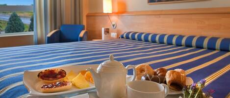 Habitación de hotel con una cama con ropa de cama a rayas azul y blanco, una mesita de noche con lámpara, y un plato de desayuno que incluye tostadas, fruta, bollería y un hervidor de agua.
