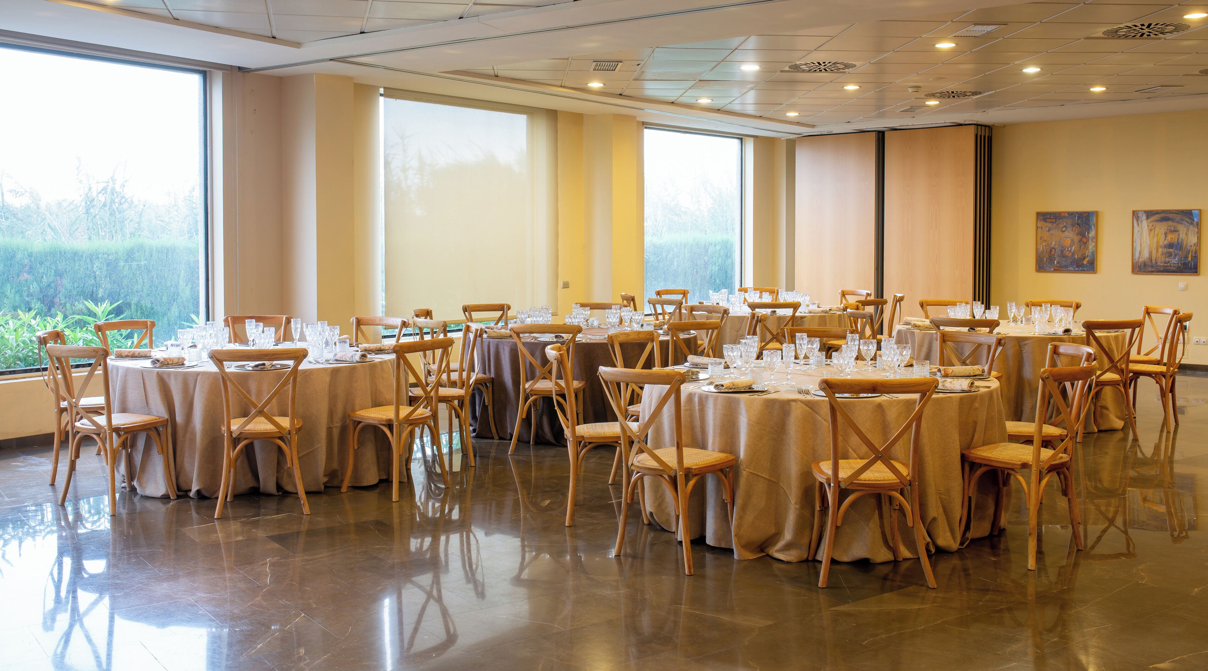 A banquet hall with round tables, wooden chairs, and large windows with a view of greenery.