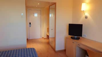 Een hotelkamer met een bed, een televisie op een houten standaard, een spiegel en een wandlamp.