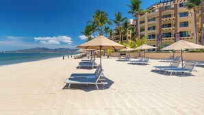 A beach with sun loungers and umbrellas, a building with balconies, and palm trees. - Villa Del Palmar Flamingos Beach Resort and Spa (Nuevo Nayarit)