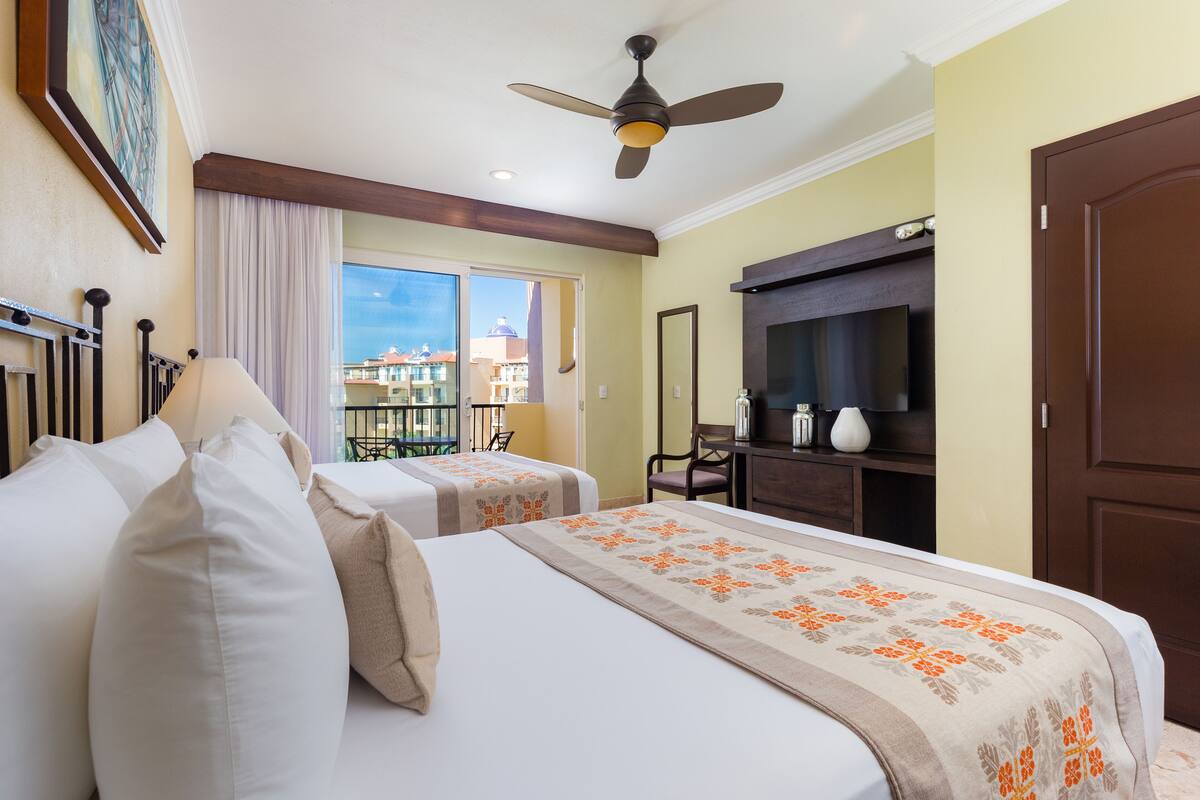 A hotel room with two beds, a ceiling fan, a TV, and a balcony with a view of buildings.