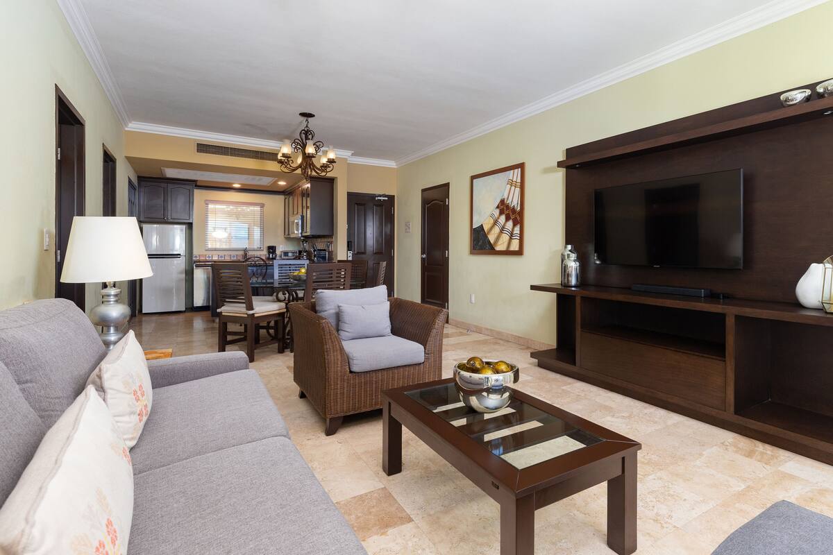 A living room with a sofa, armchair, coffee table, TV, and kitchen in the background.