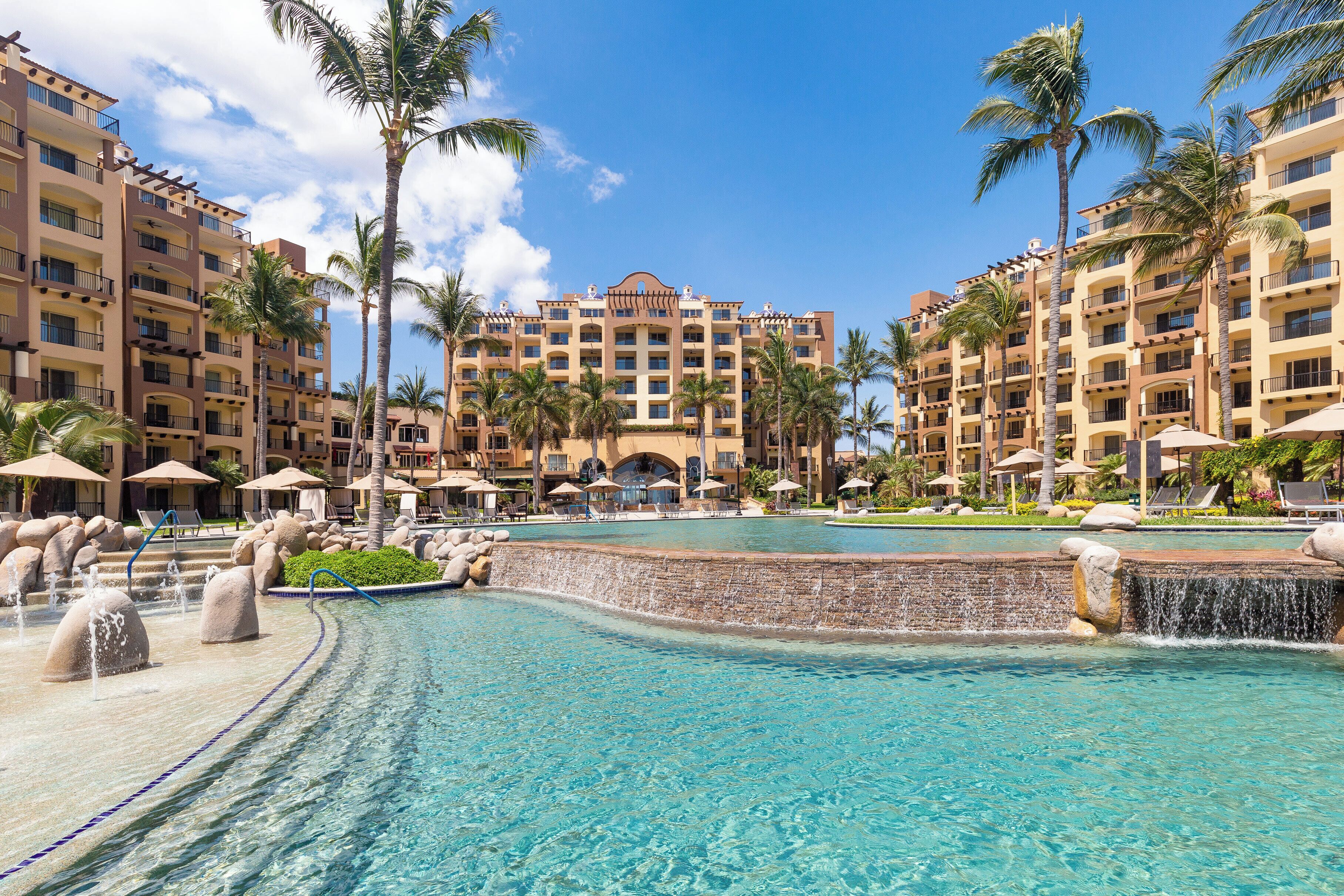 A resort with a swimming pool, palm trees, and a multi-story building with balconies.
