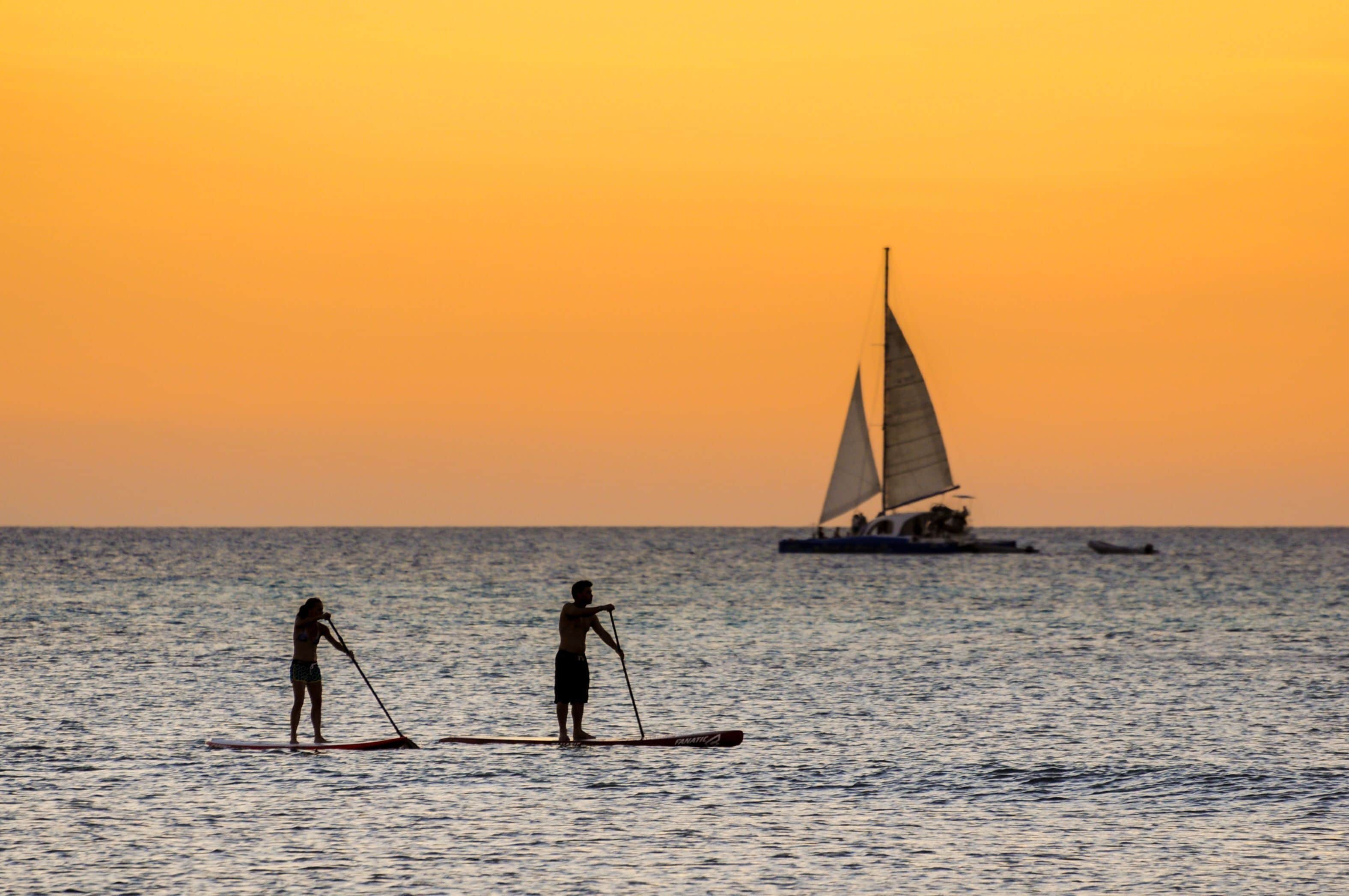 Two people paddleboarding on calm water with a sailboat in the background during sunset.