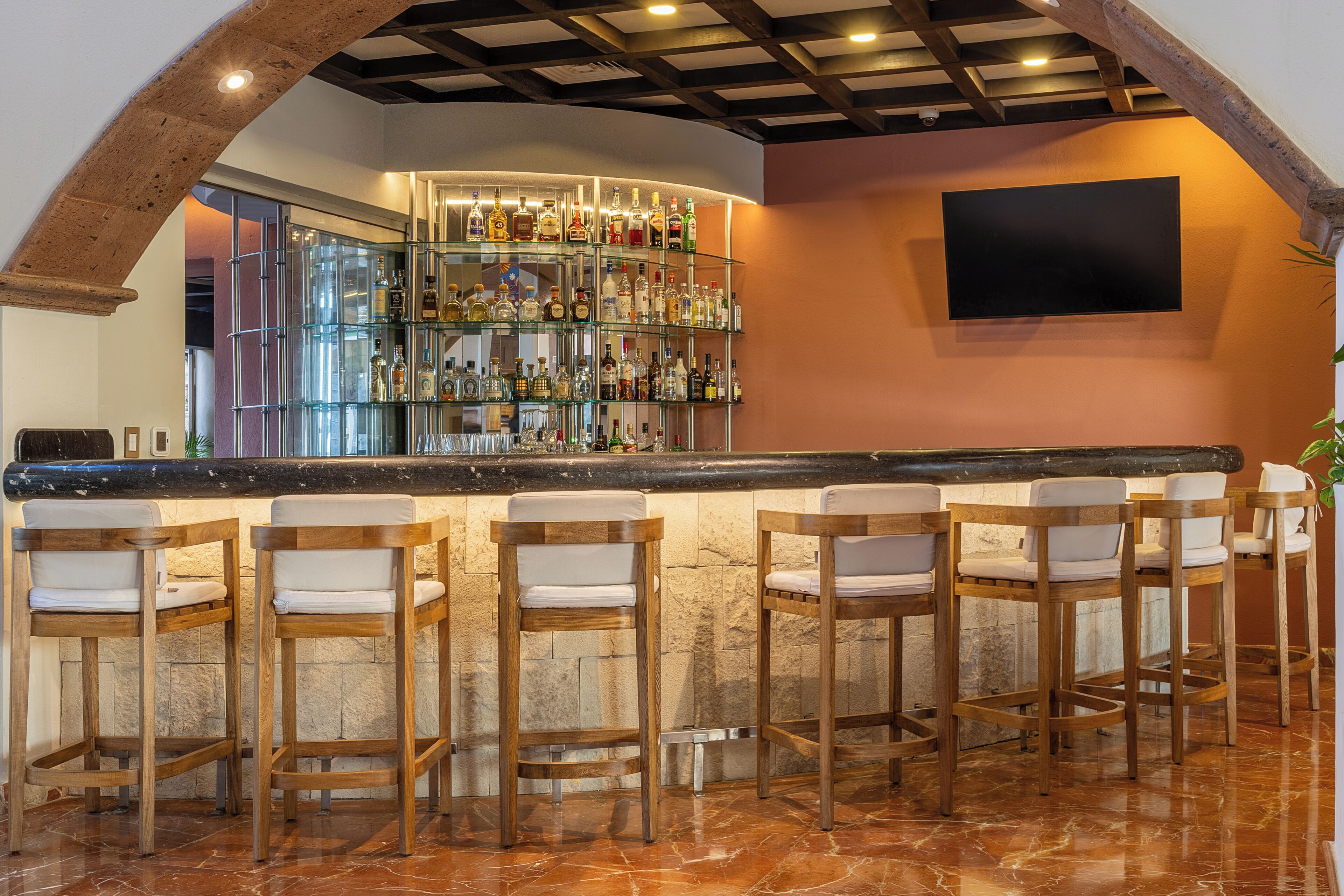 A bar area with a marble countertop, wooden bar stools, and a glass shelving unit displaying various bottles.