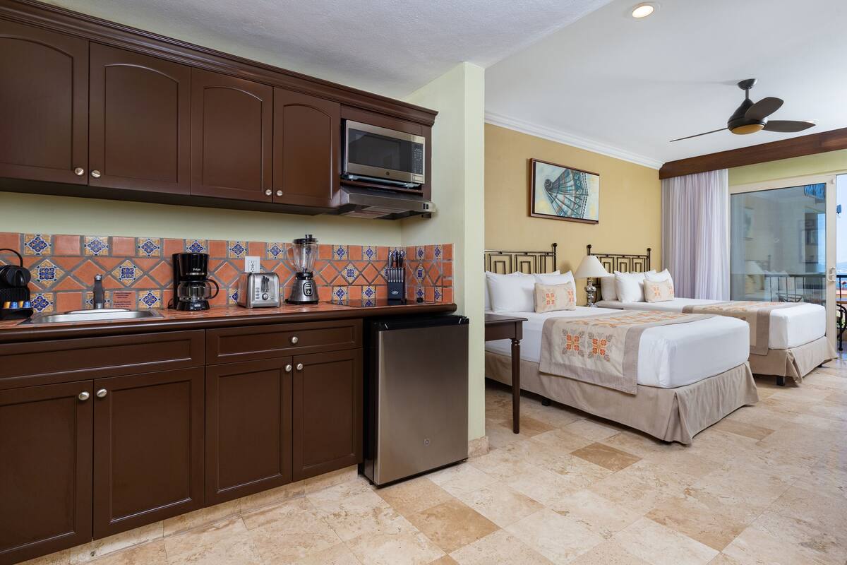 A hotel room with a kitchenette, a bed with two pillows, a bedside table with a lamp, and a view of the outdoors.