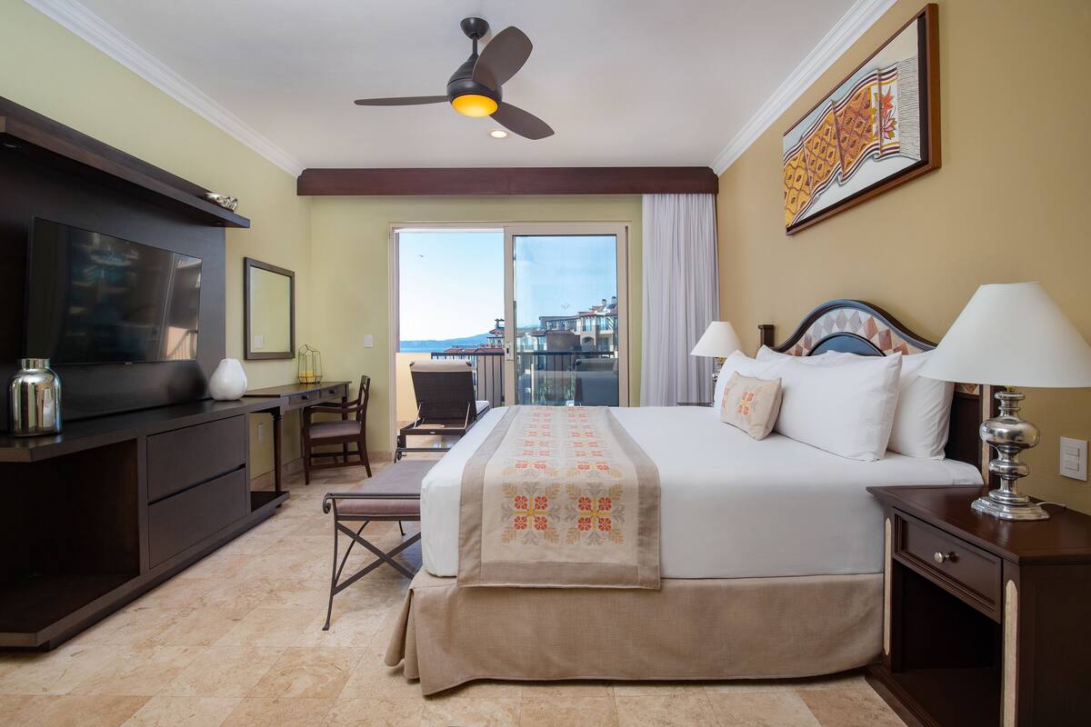 A hotel room with a bed, a desk, a chair, a TV, and a balcony with a view.