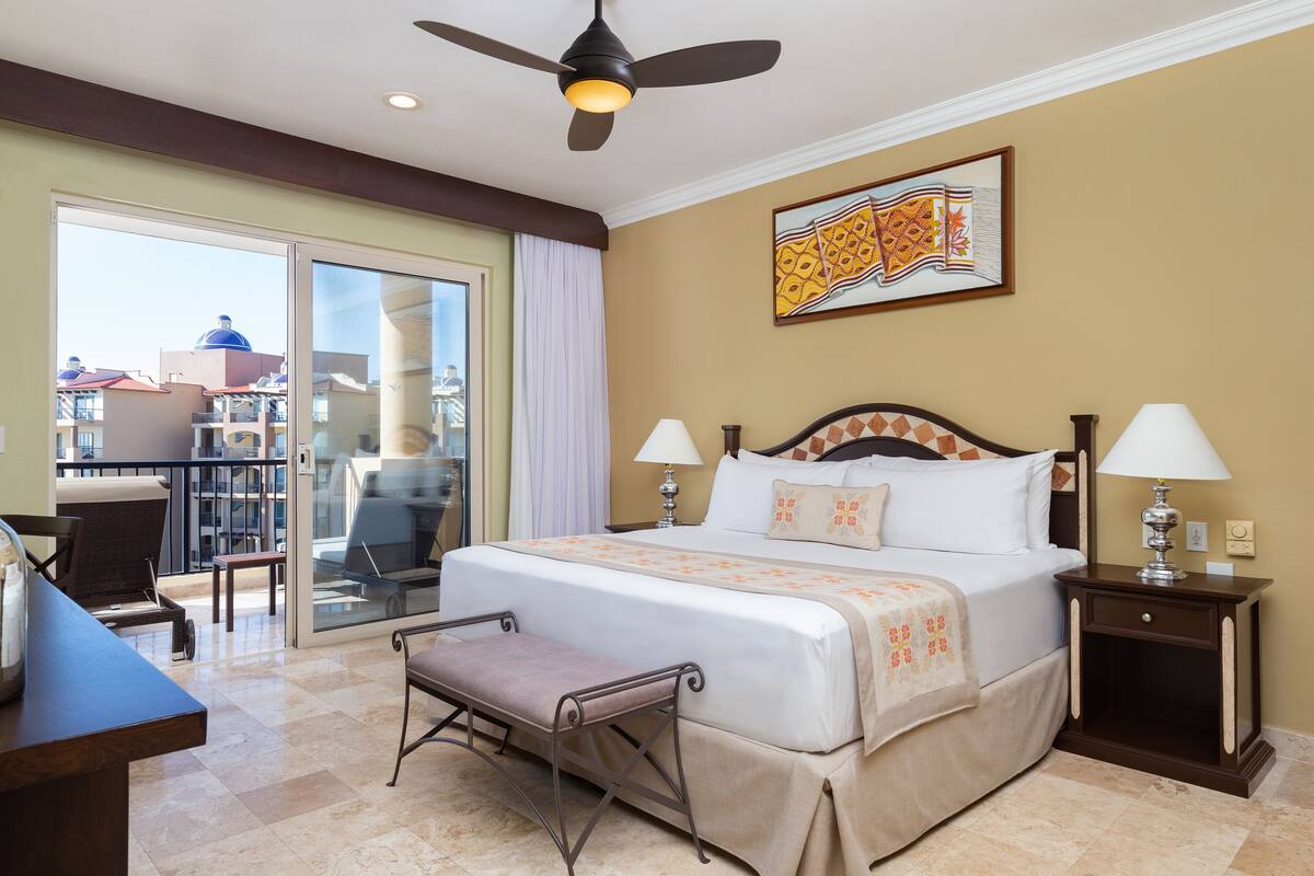 A hotel room with a large bed, two bedside lamps, a ceiling fan, a balcony with a view, and a desk with a chair.