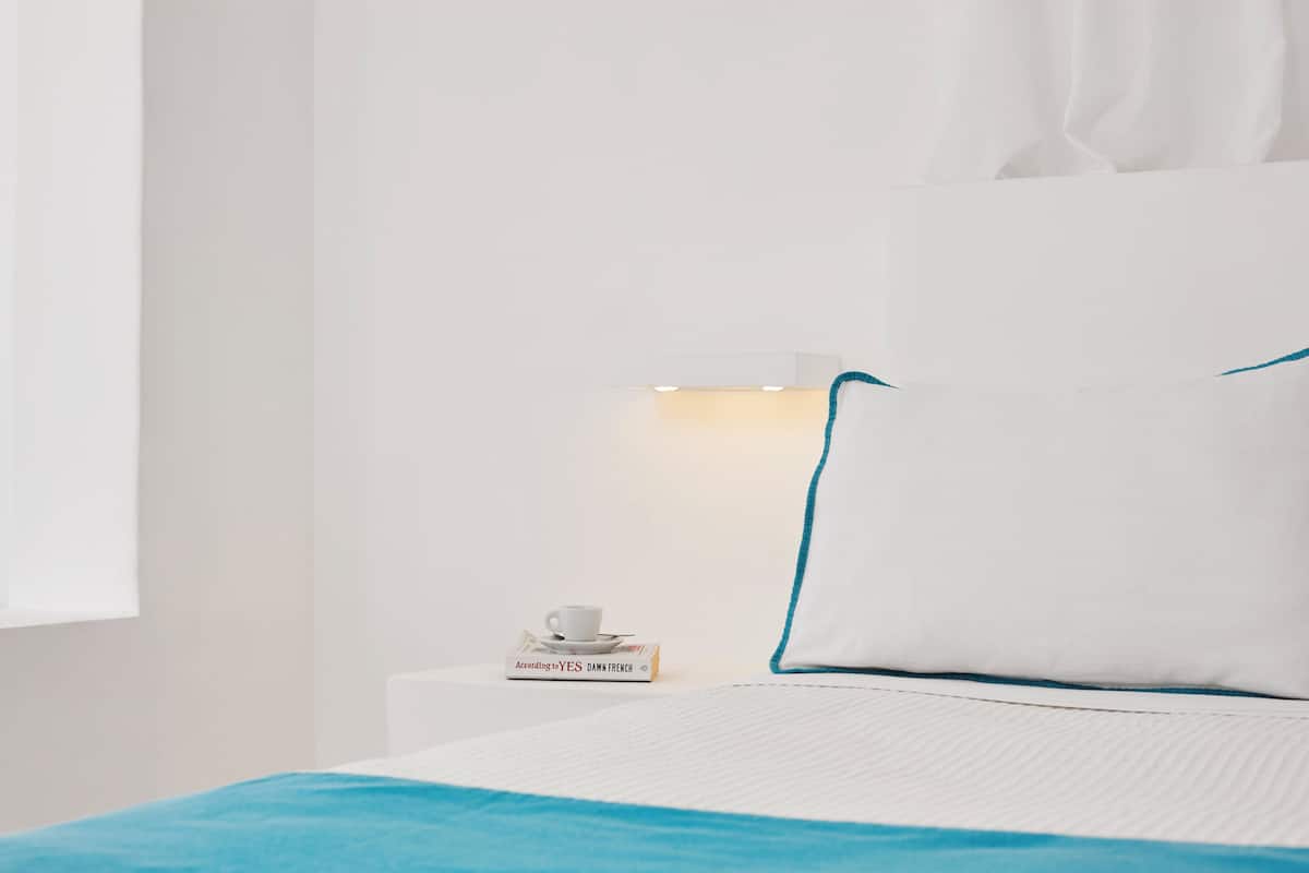 A neatly made bed with a white and blue bedspread, a white pillow with a blue trim, and a bedside table with a cup and a book titled 'According to YES, DAWN FRENCH'.