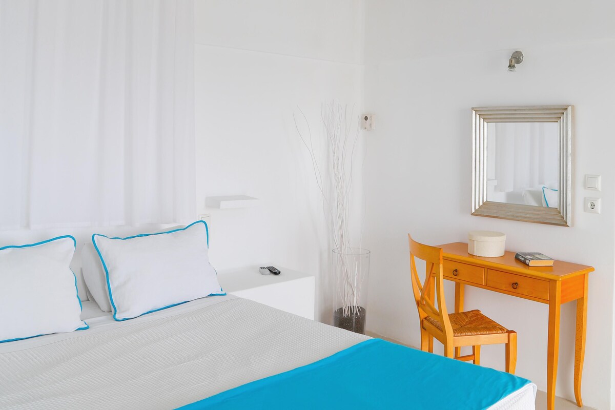 A hotel room with a bed, a desk, a chair, a mirror, and a vase with branches.