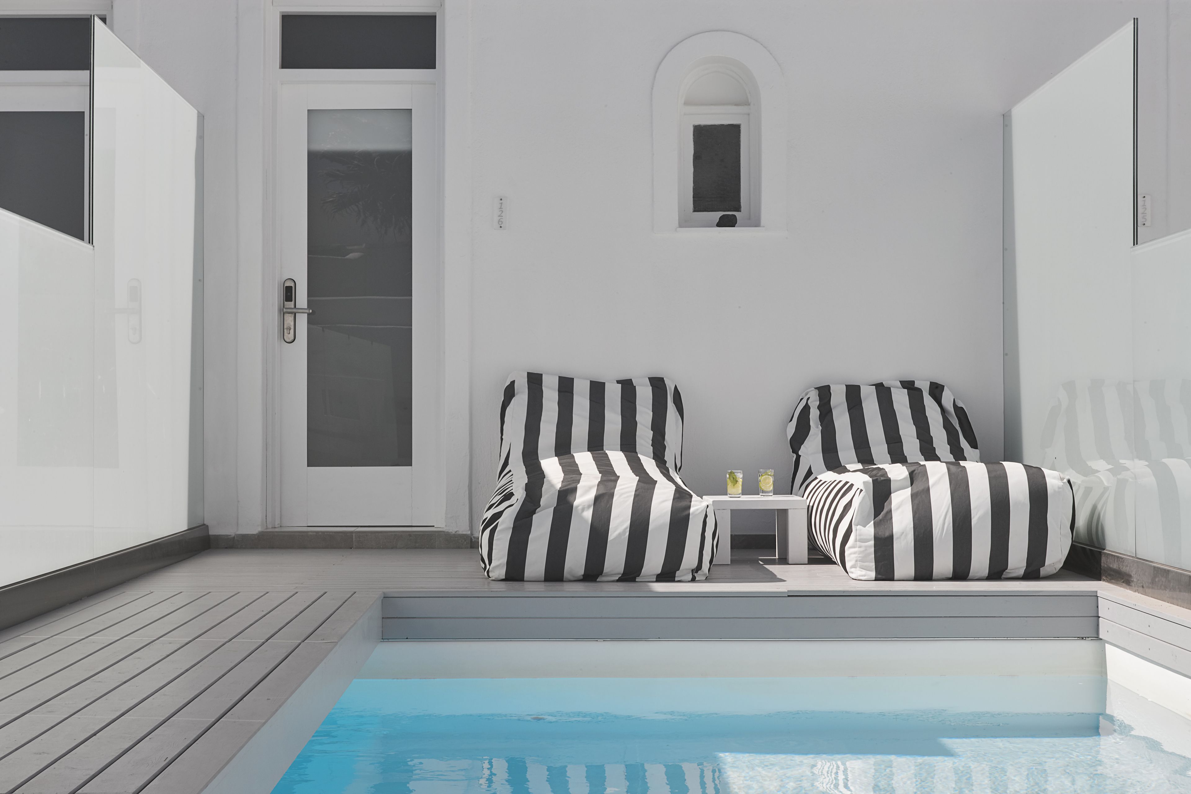A modern outdoor pool area with a wooden deck, two striped lounge chairs, a small table with drinks, and a door.