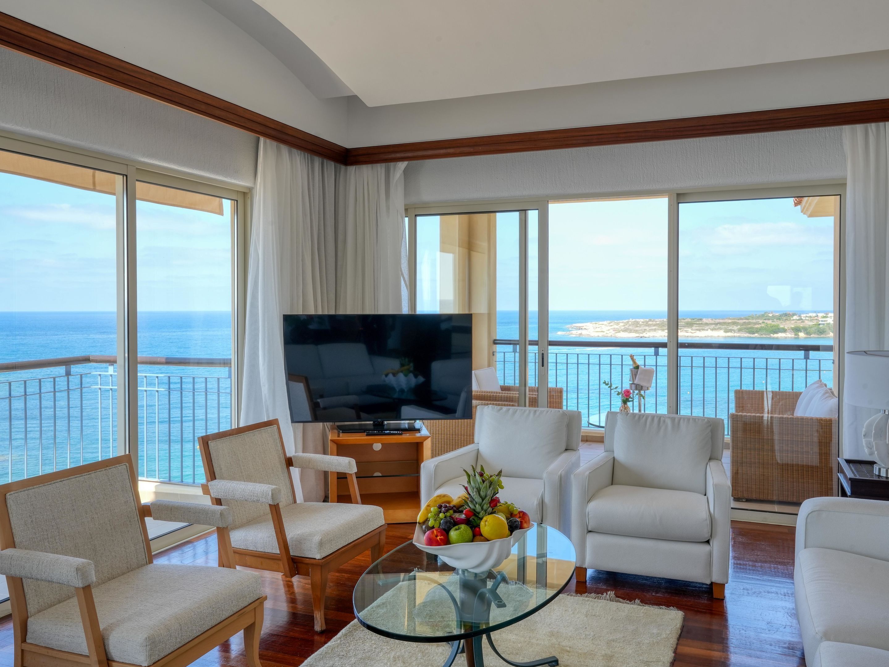 A hotel room with a balcony, a flat-screen TV, a glass coffee table with fruit, and a view of the sea.