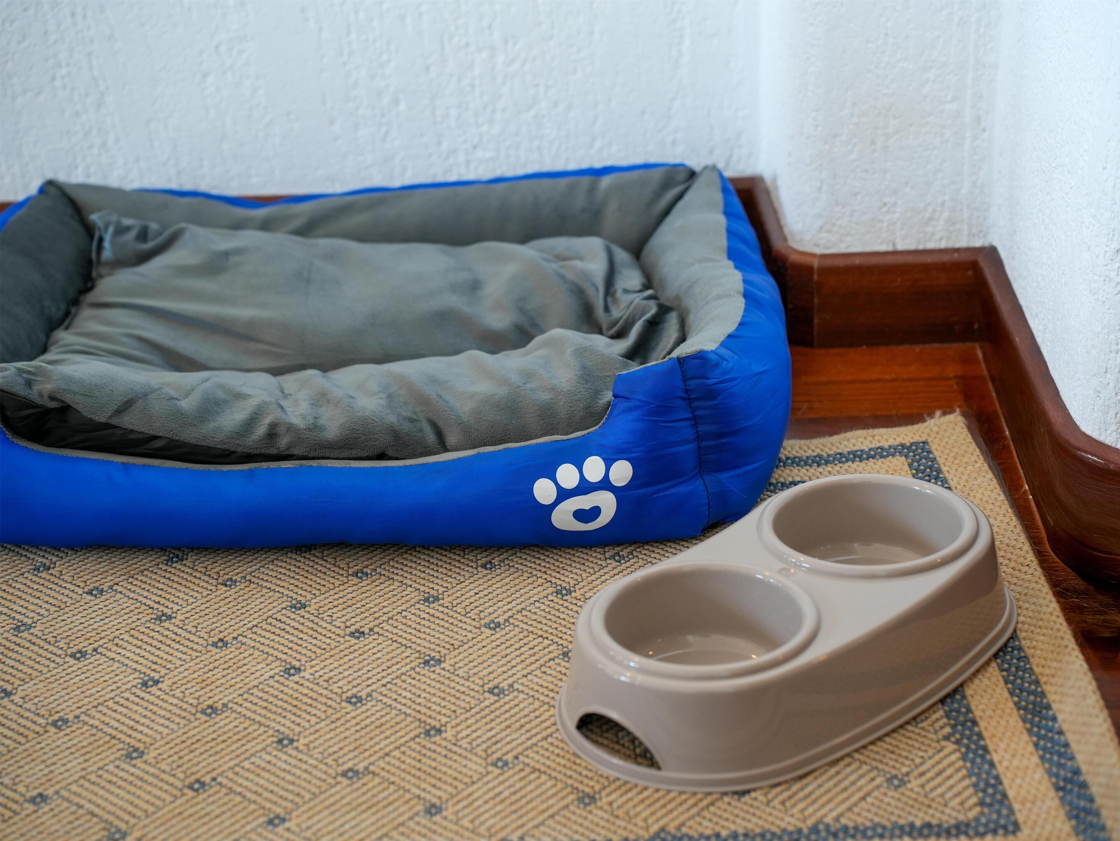 Un panier pour animaux de compagnie orné d’un motif représentant des empreintes de pattes, accompagné d’une paire de gamelles posées sur un tapis tissé.