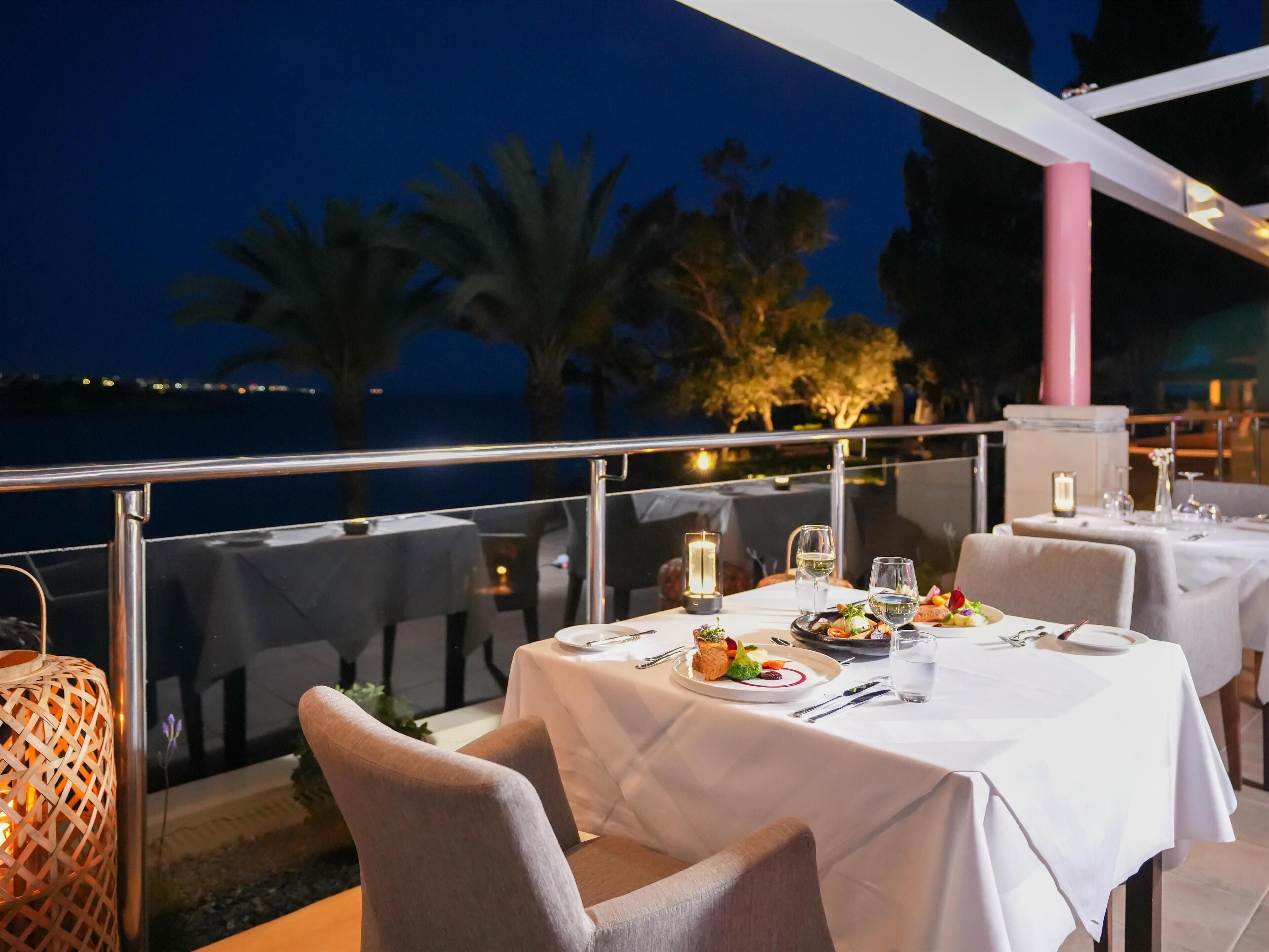 An outdoor dining area with tables set for two, overlooking a body of water at night.