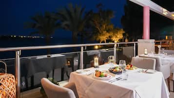 An outdoor dining area with tables set for two, overlooking a body of water at night.