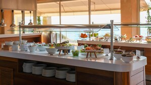 Disposição de buffet com diversos alimentos e recipientes, uma vitrine de vidro e vista para o exterior através de grandes janelas.