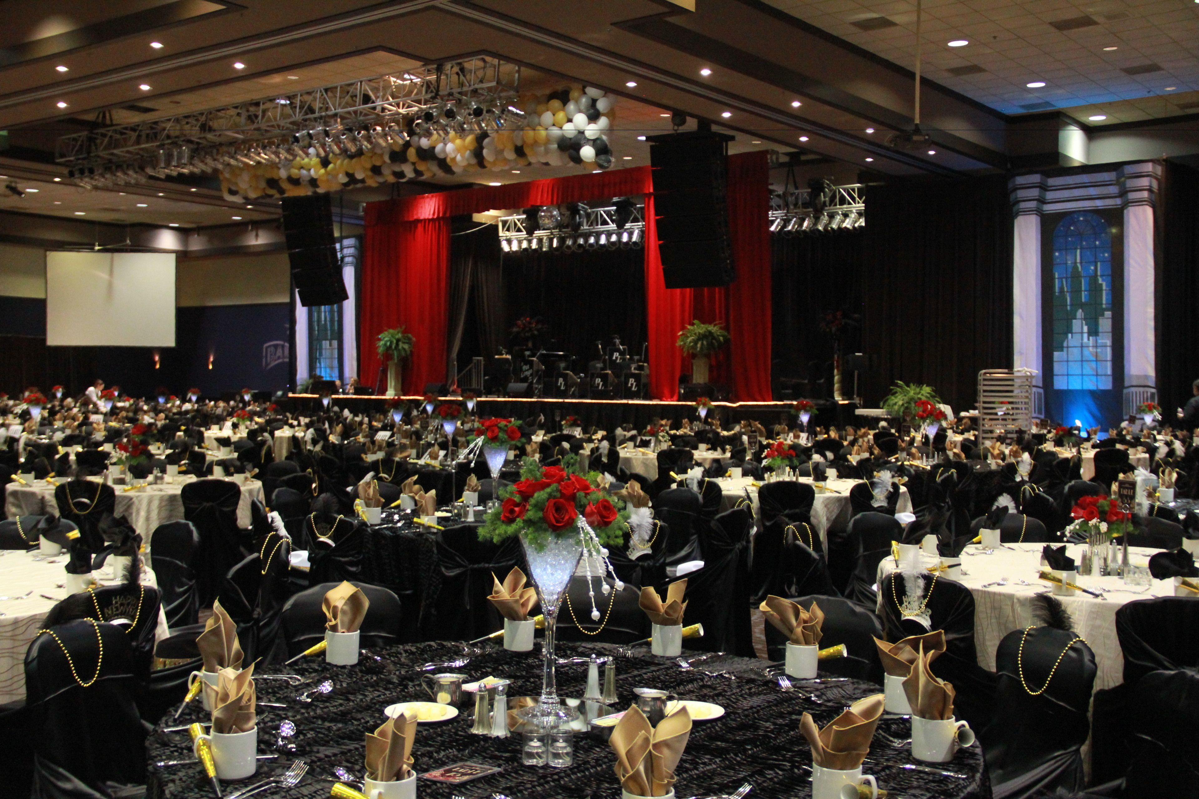 Un salón de banquetes con mesas redondas dispuestas para un evento formal, con un escenario que incluye instrumentos musicales y una amplia audiencia.