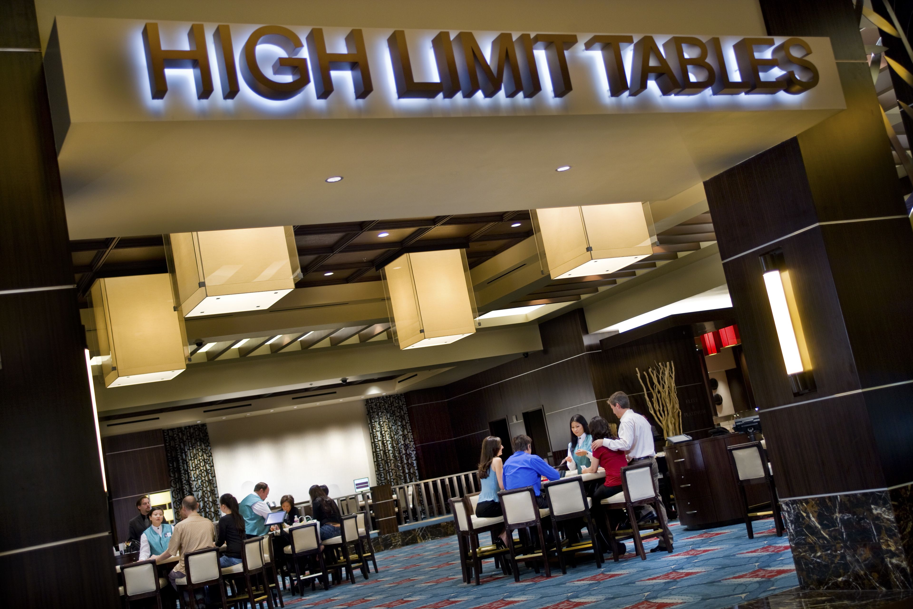 Ein Konferenzraum mit Tischen und Stühlen, einem Schild mit der Aufschrift 'HIGH LIMIT TABLES' und einer Gruppe von Menschen, die an einer Besprechung teilnehmen.