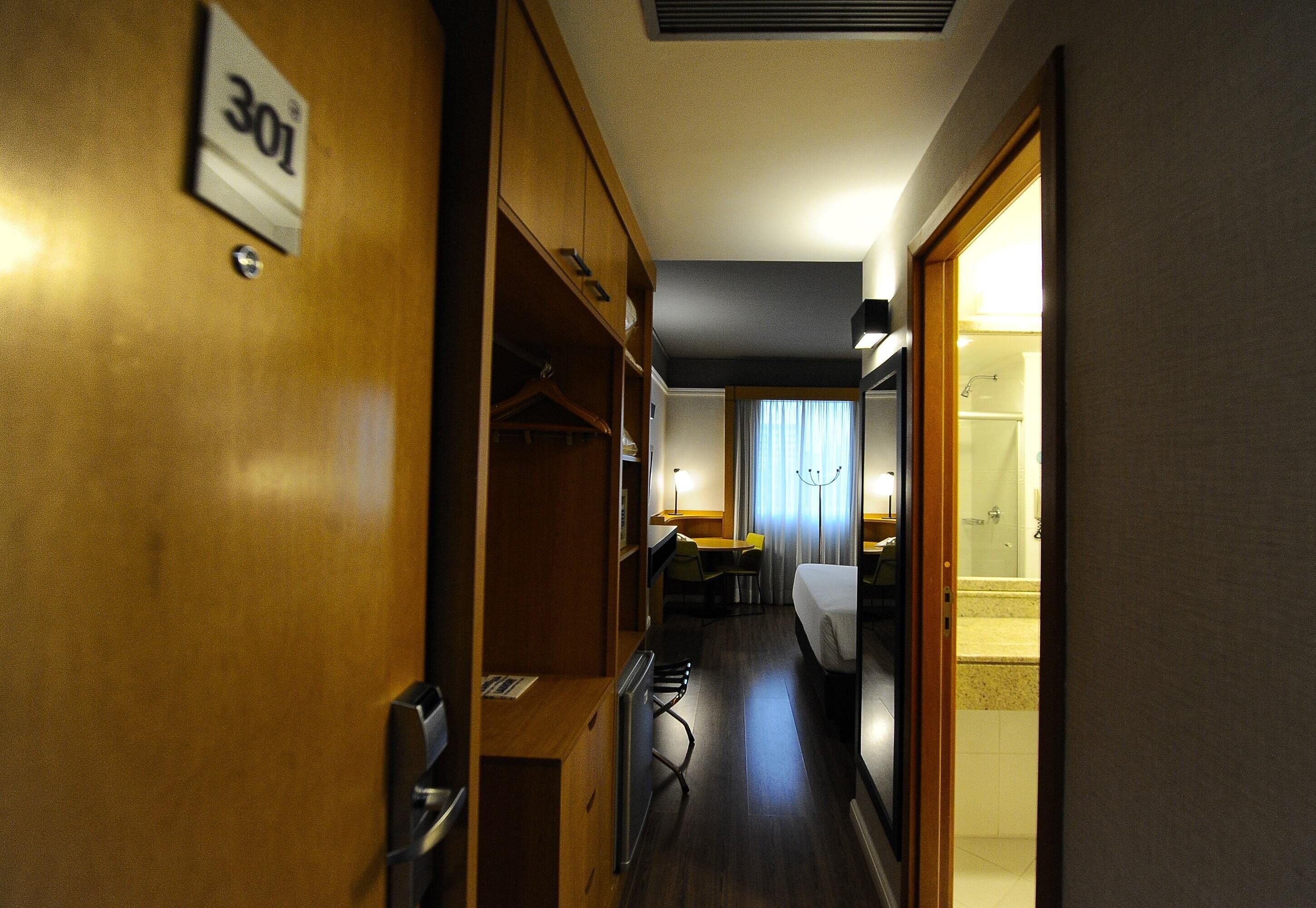 Ein Hotelkorridor mit einem Bett, einem Schreibtisch und einem durch eine offene Tür einsehbaren Badezimmer.