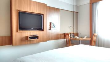 Una habitación de hotel con una cama grande, un televisor montado en la pared, un escritorio con una silla y una ventana con cortinas.
