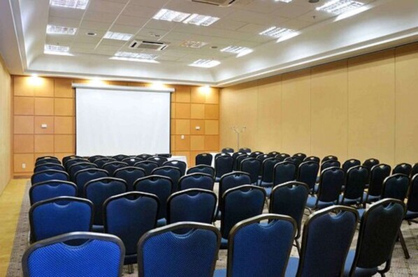 A conference room with rows of blue chairs, a white projection screen, and a wooden panel wall. - Bristol Santo André Hotel (Santo André)