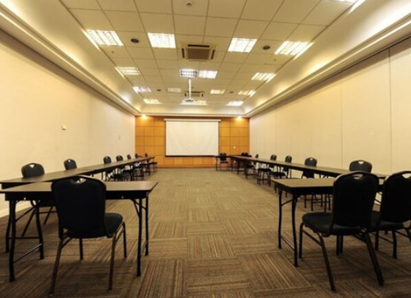 Un salón de conferencias con una pantalla grande al frente, varias mesas con sillas y piso alfombrado.