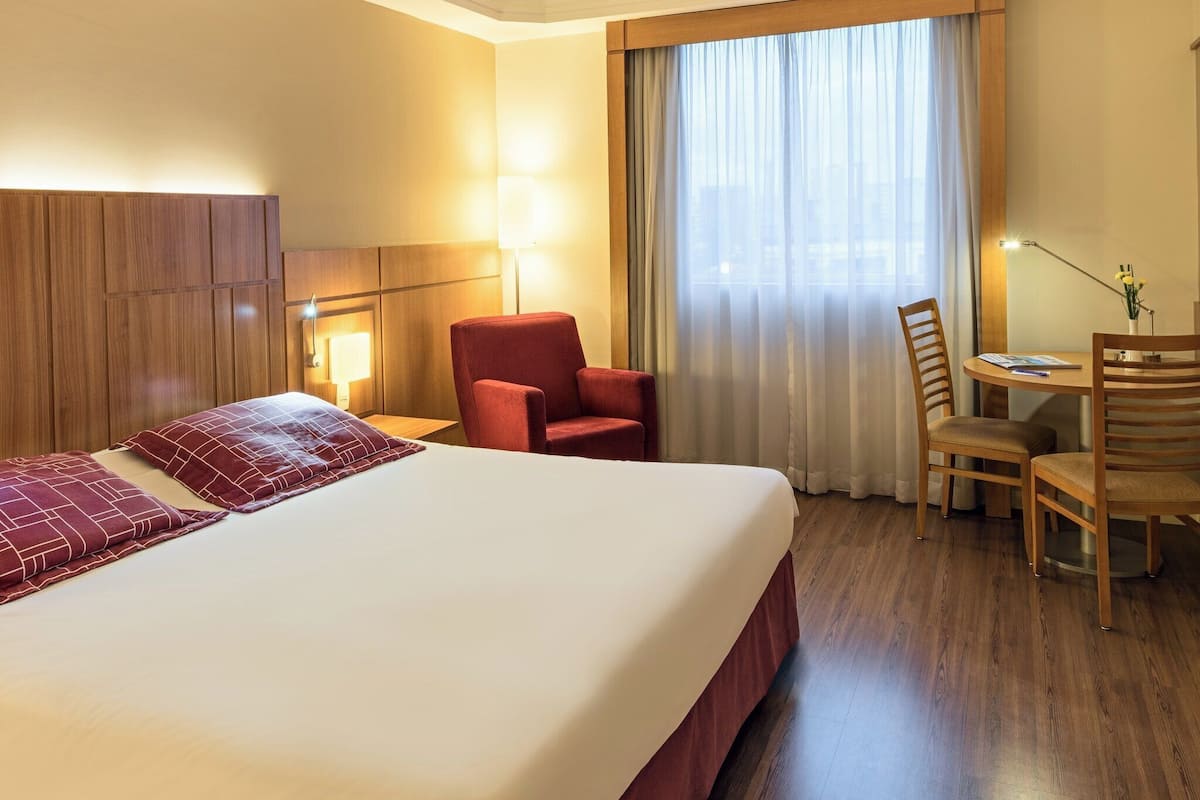 Une chambre d’hôtel avec un grand lit, un bureau, deux chaises, une lampe et une vue sur la ville par la fenêtre.