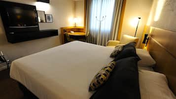 A hotel room with a large bed, a flat-screen TV mounted on the wall, a desk with a chair, and a lamp.