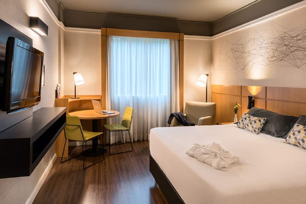 Une chambre d’hôtel comprenant un lit, un bureau avec une chaise, un téléviseur, une fenêtre avec des rideaux et un mur orné d’un motif géométrique.