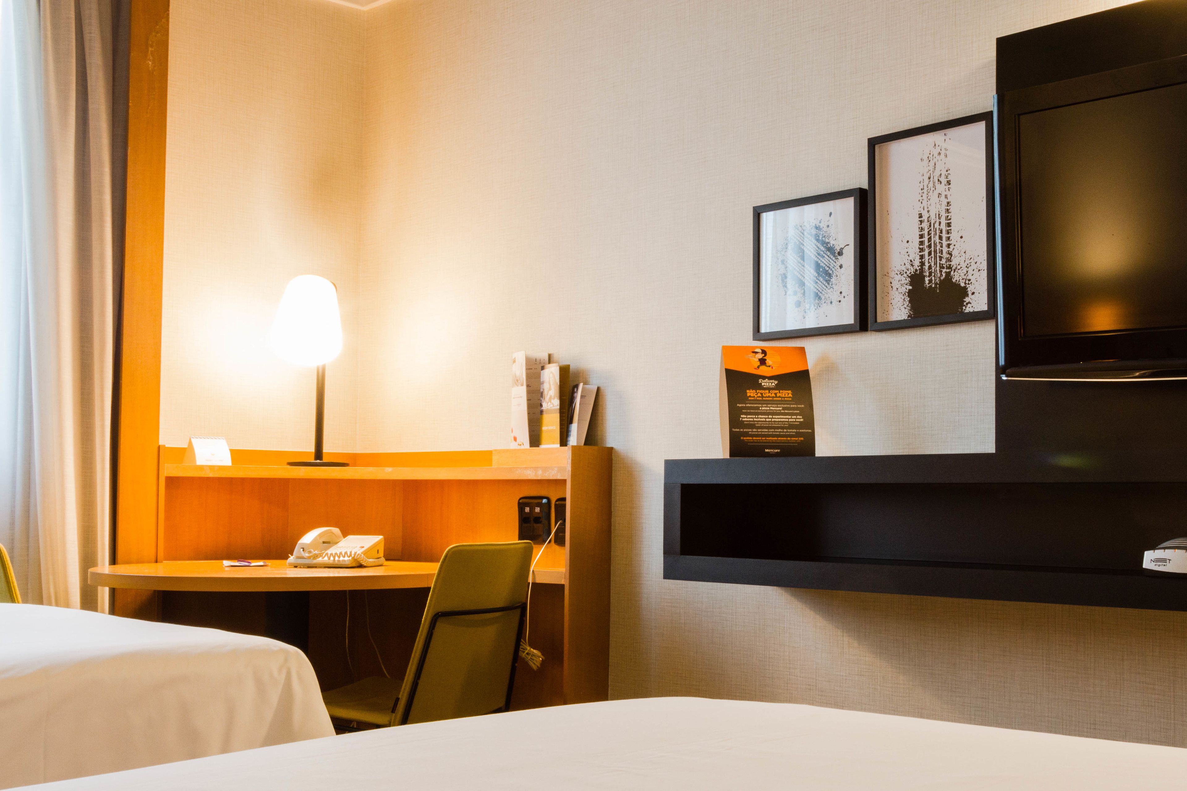 A hotel room with two beds, a desk with a lamp, a telephone, a TV, and framed pictures on the wall.