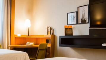 A hotel room with two beds, a desk with a lamp, a telephone, a TV, and framed pictures on the wall.