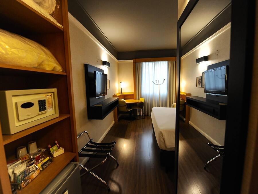 Ein Hotelzimmer mit einem Bett, einem Schreibtisch, einem Sessel, einem Fernseher, einem Spiegel und einem Regal mit Pflegeprodukten.