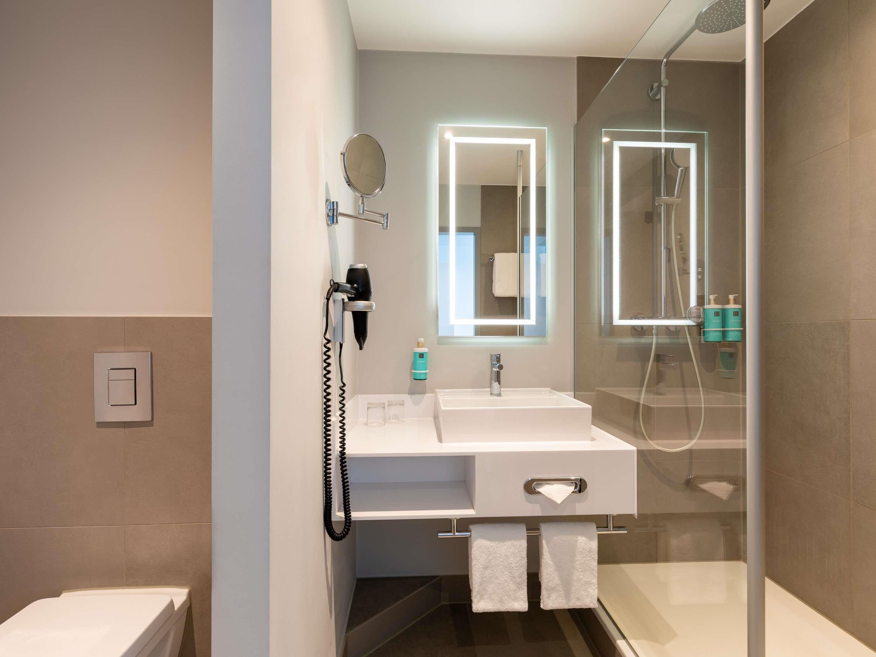 Ein modernes Badezimmer mit einer gläsernen Duschkabine, einem weißen Waschbecken mit Spiegel und einem an der Wand befestigten Föhn.