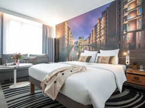 Une chambre d’hôtel avec un grand lit, un bureau agrémenté d’un vase de fleurs et une fresque représentant un paysage urbain sur le mur.