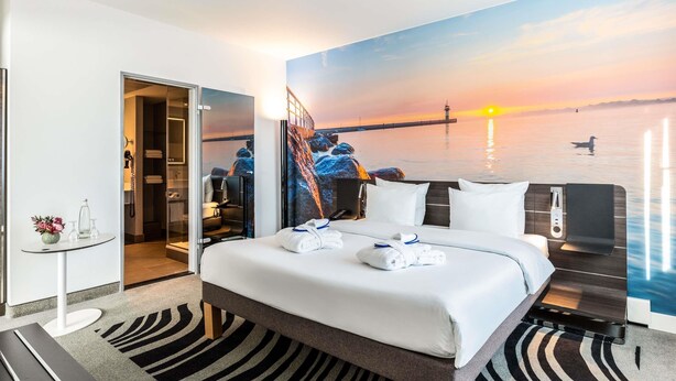 Une chambre d’hôtel comprenant un lit, un bureau, une chaise, une petite table avec un vase et un tableau représentant un coucher de soleil sur l’eau.