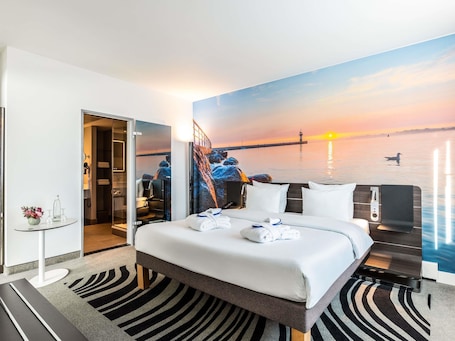 A hotel room with a bed, a desk, a chair, a small table with a vase, and a painting of a sunset over water.