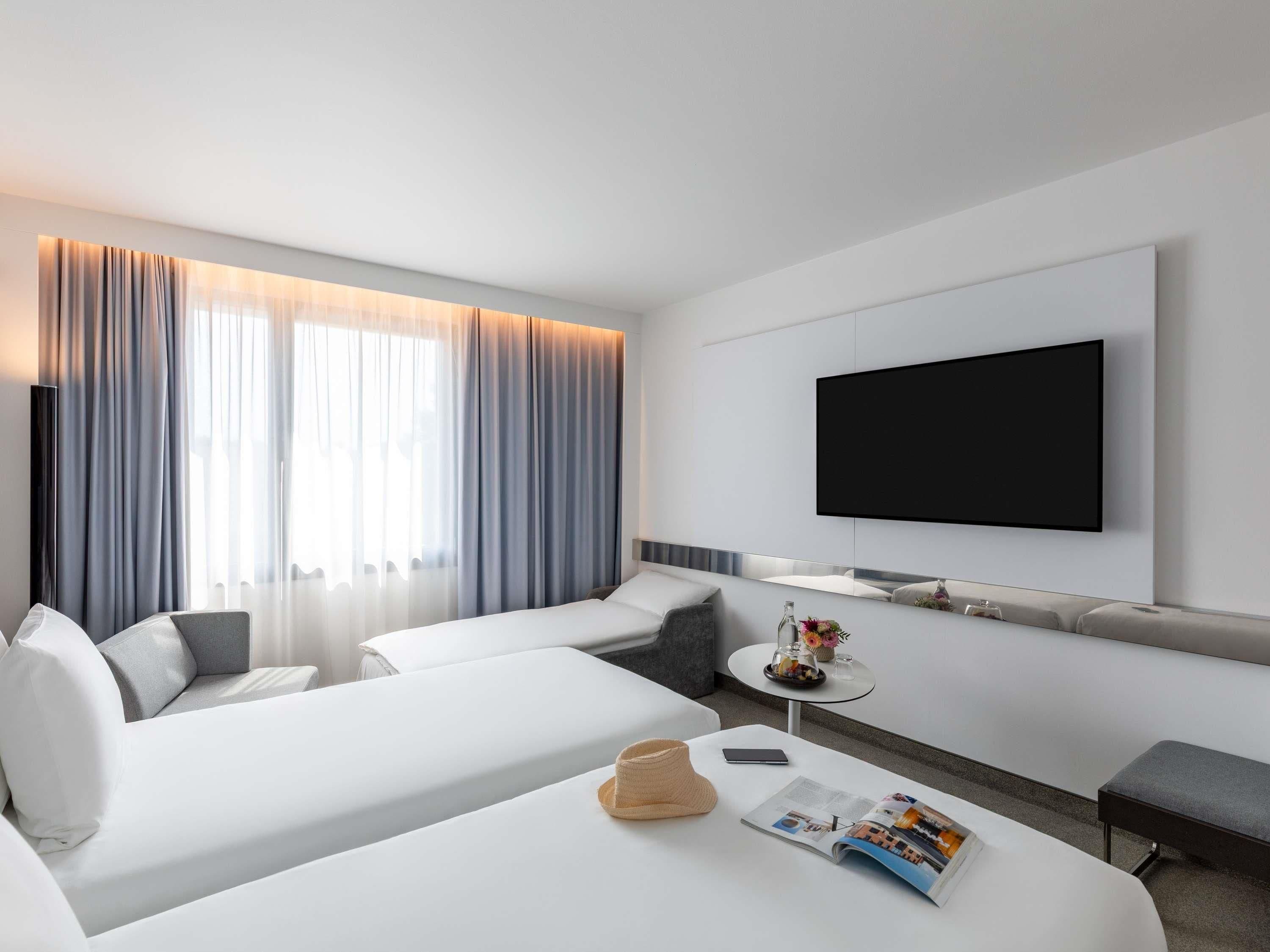 A hotel room with two beds, a TV, a small table with a magazine, and a window with curtains.