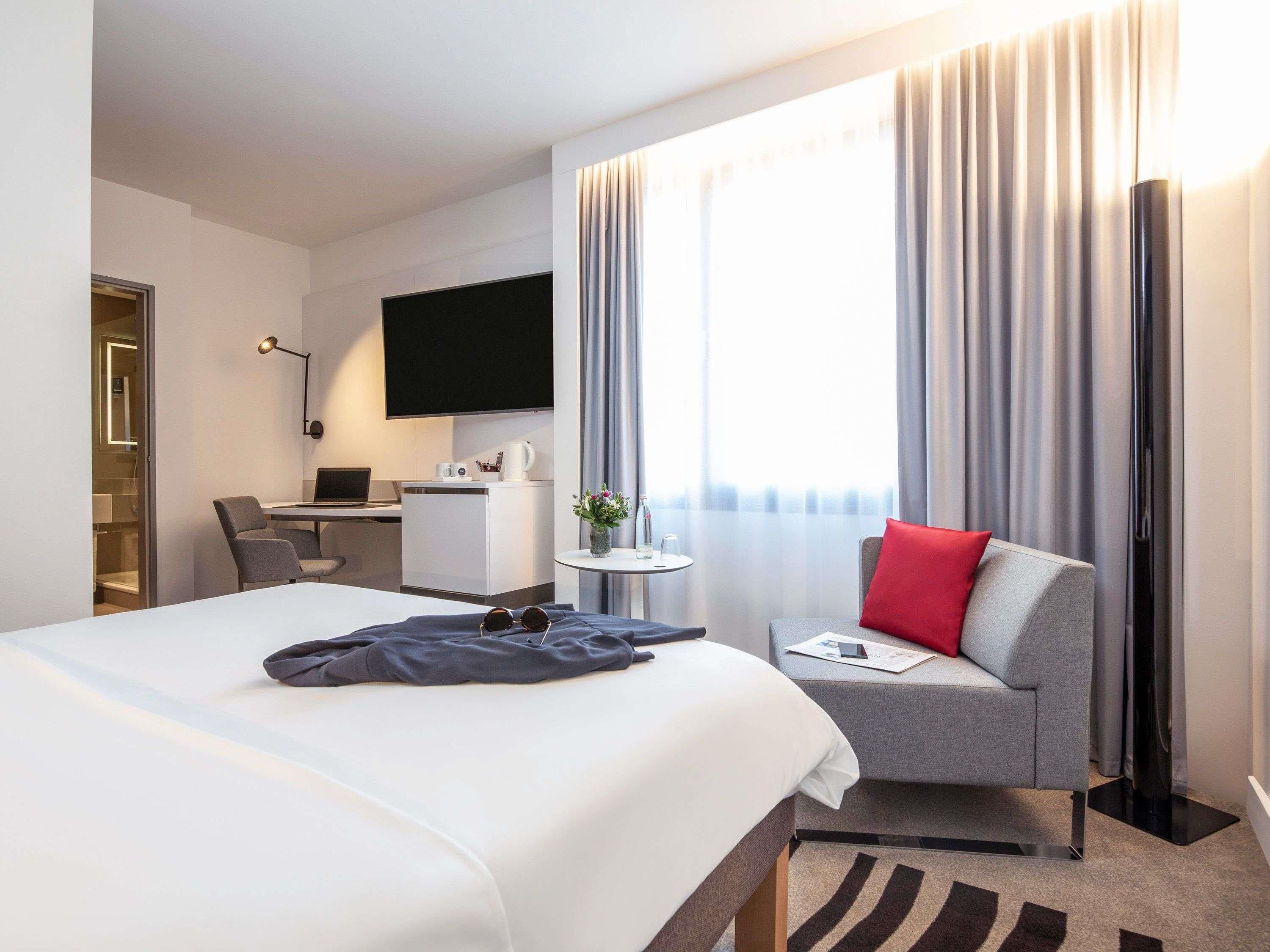 Habitación de hotel con una cama, un escritorio con computadora, una silla, un televisor y una ventana con cortinas.