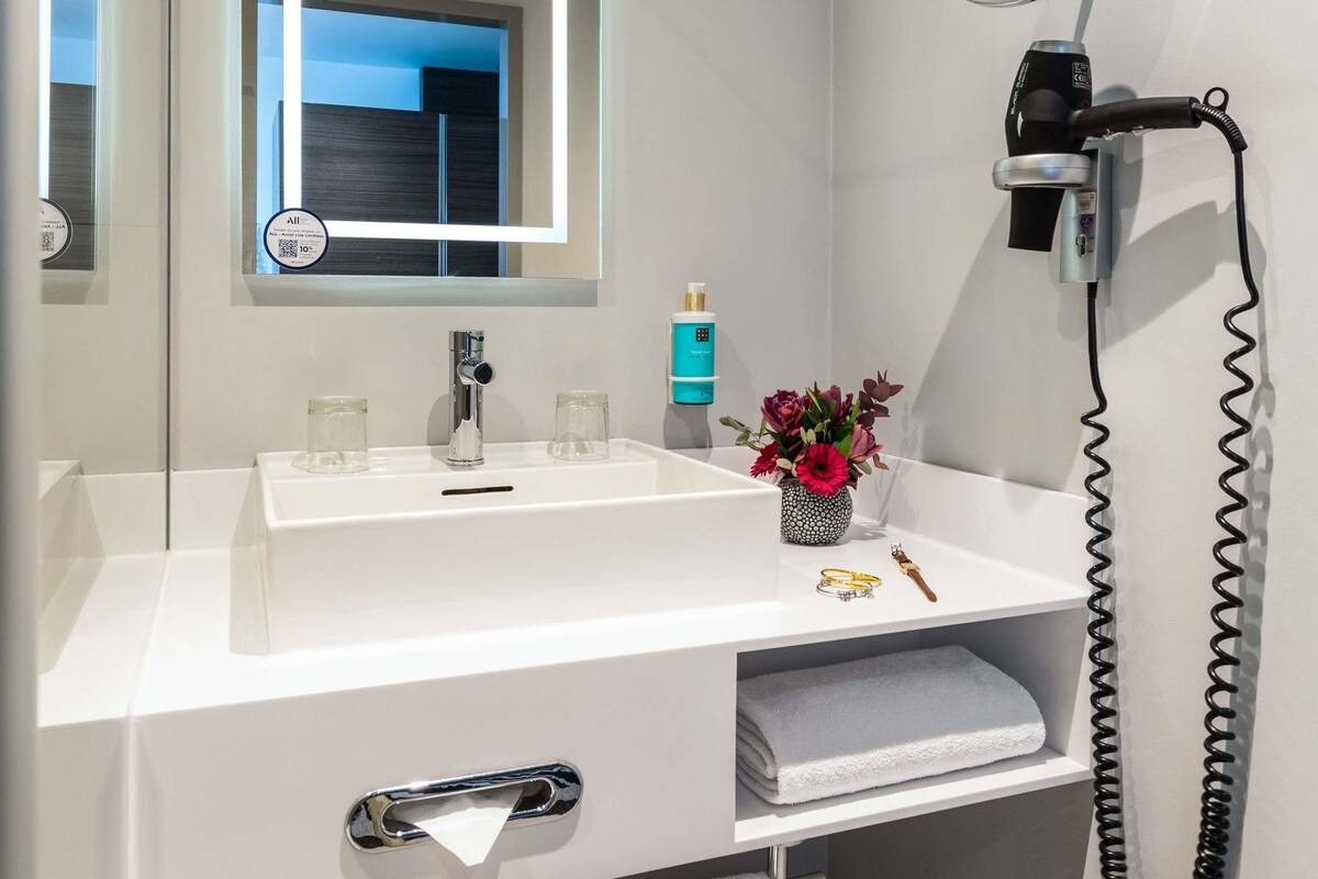 Une salle de bain moderne avec un lavabo blanc, un sèche-cheveux, un petit vase avec des fleurs et un porte-serviettes.