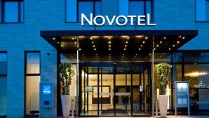 L’extérieur d’un hôtel Novotel, avec une entrée en verre, des plantes en pot et une façade bleue.