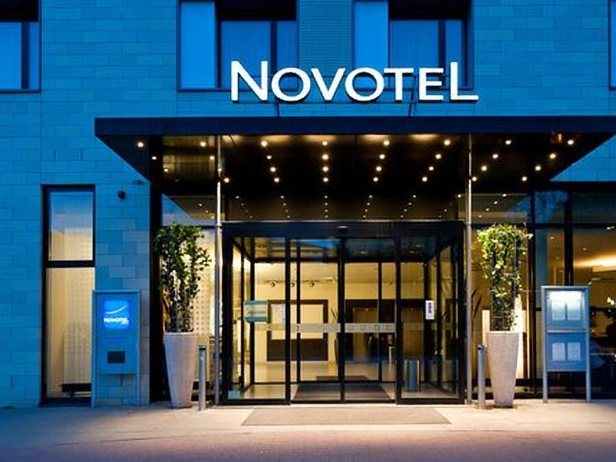 Exterior de un hotel Novotel con entrada de vidrio, plantas en macetas y fachada azul.