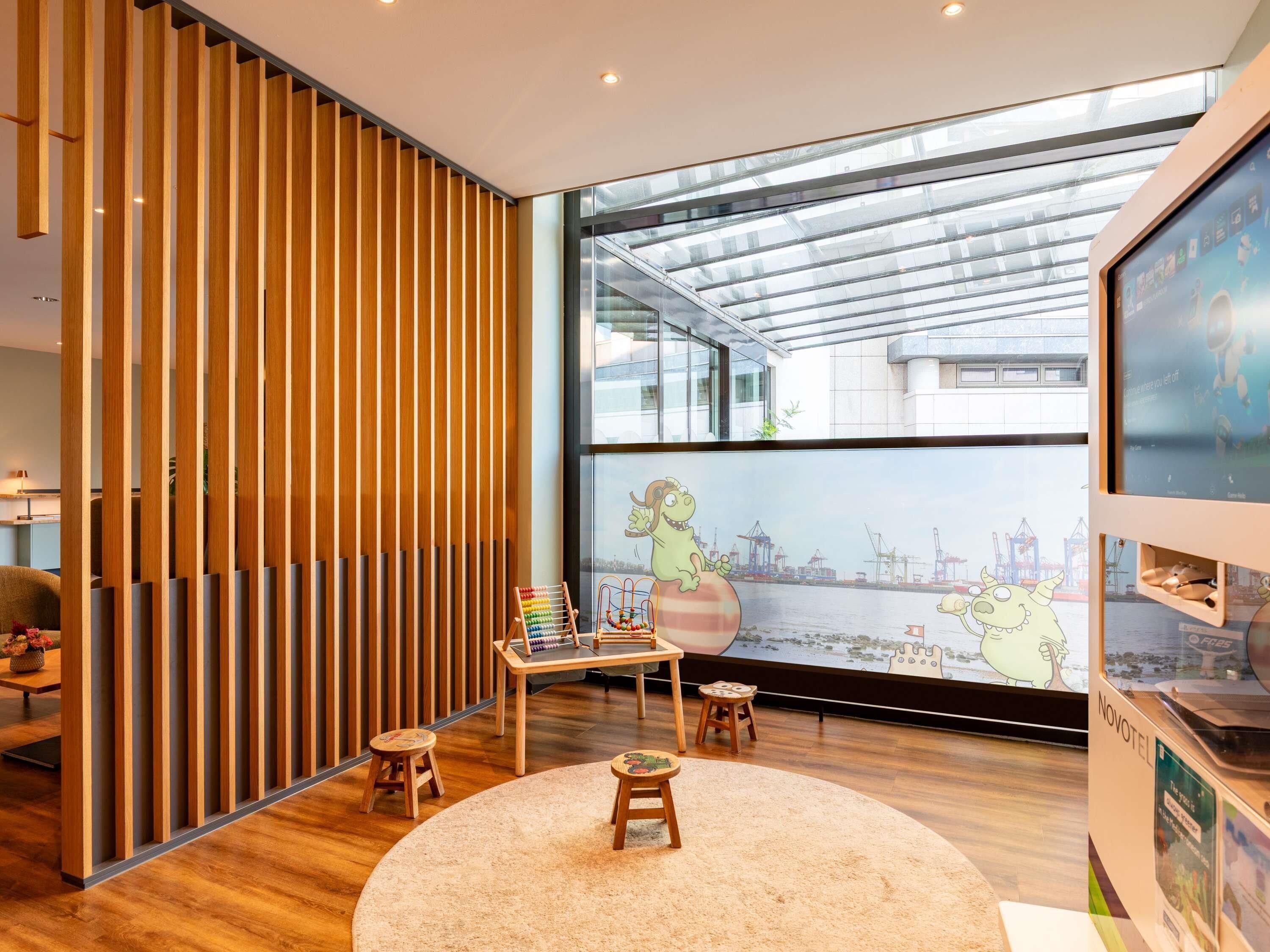 A modern interior with a wooden slat partition, a large window with a view of a port, and a children's play area with a colorful abacus and a cartoon character.
