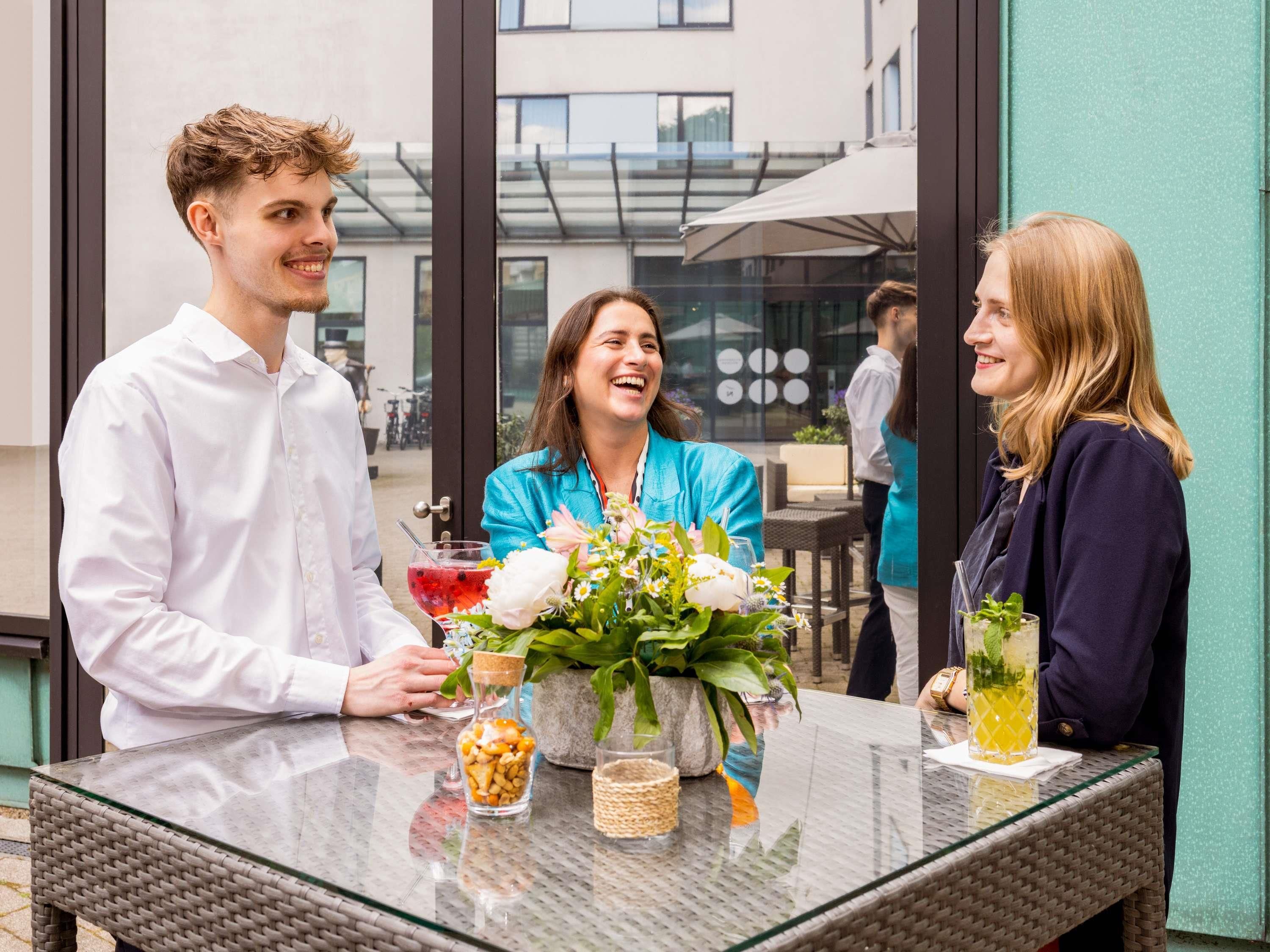 A glass table with a vase of flowers, a glass of drink, and a container of snacks.