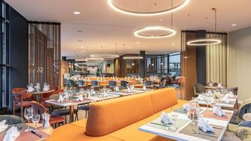 Un restaurante moderno con varias zonas de comedor, incluyendo un área central con asientos tipo banca naranja y mesas individuales.