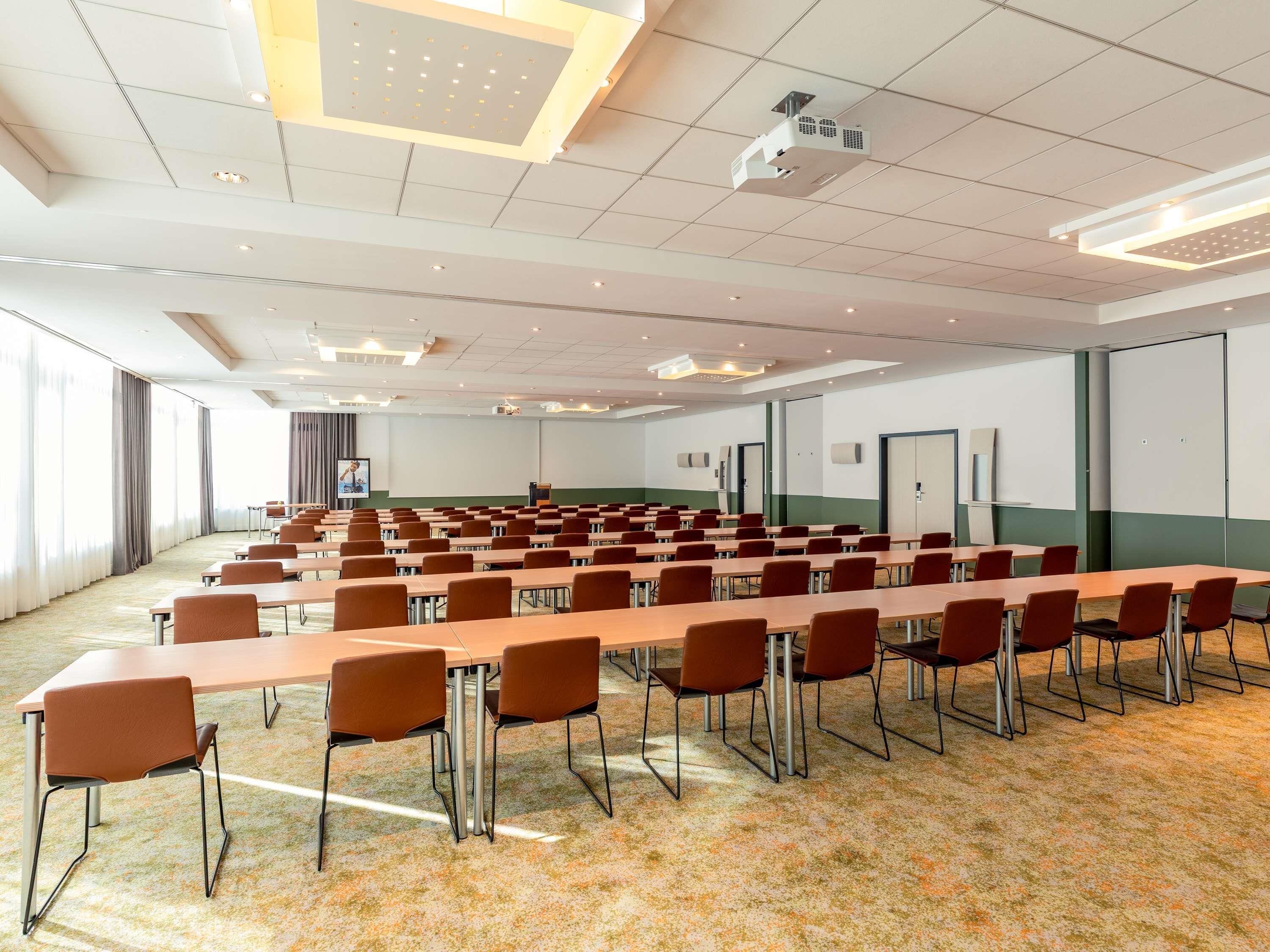 A conference room with rows of tables and chairs, a projector, and a large screen.